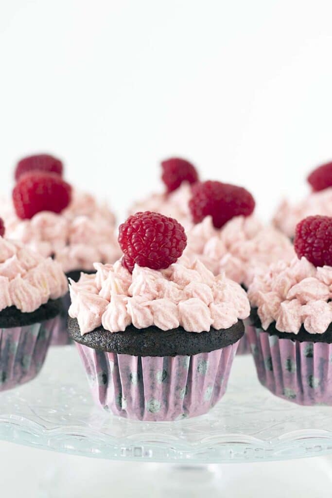 vegan chocolate cupcakes with pink frosting and raspberries on glass cake stand