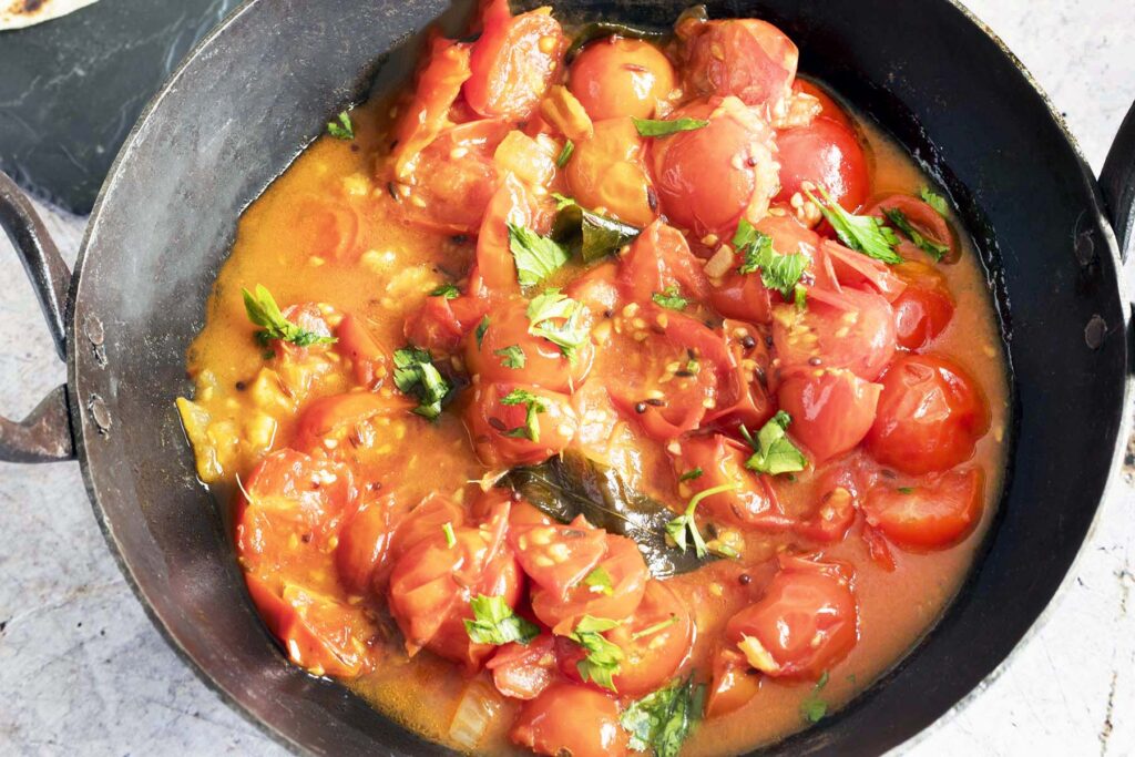 fresh tomato curry in bowl with rice, pan and breads in background