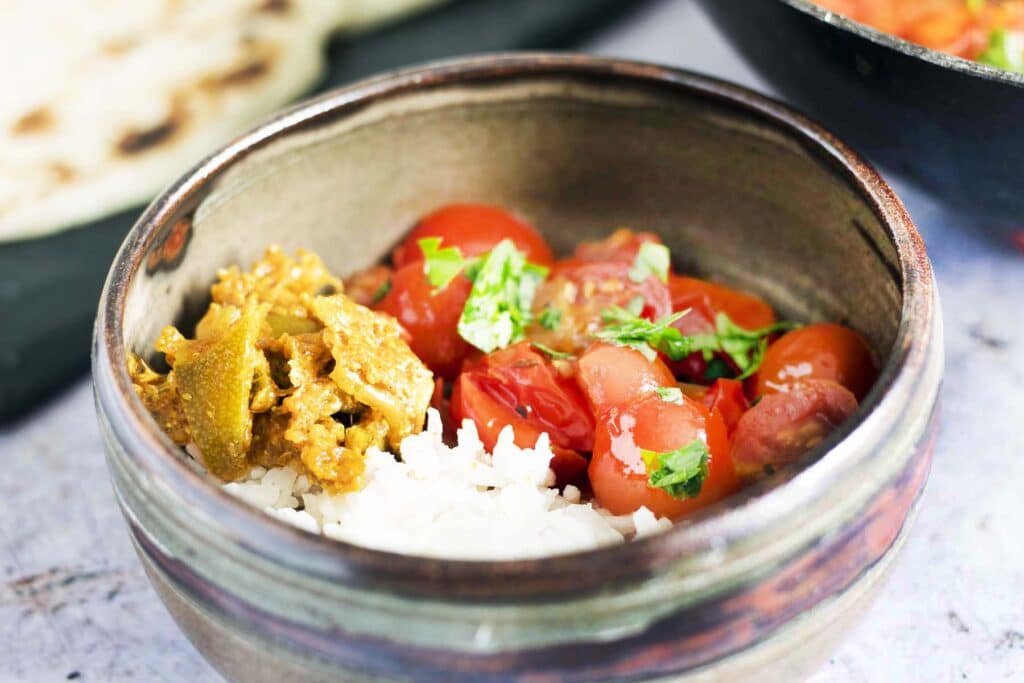 fresh tomato curry in bowl with rice, pan and breads in background